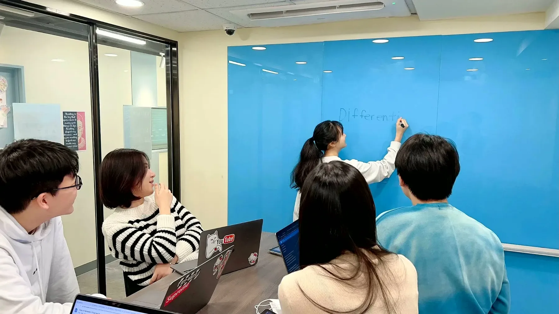 High school students collaborating around a table while one writes on a blue glass whiteboard wall.